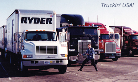 Male in blue plaid shirt posing with white Ryder truck and line of 18 wheelers in the background