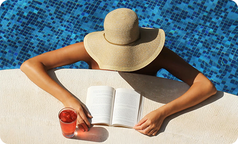 Video still of overhead view of woman with floppy sun hat and tanned arms reading a book by the side of a swimming pool