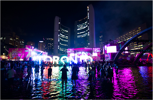 Photo at night with people in silhouette, colourful water reflections and illuminated Toronto sign at Nathan Phillips square